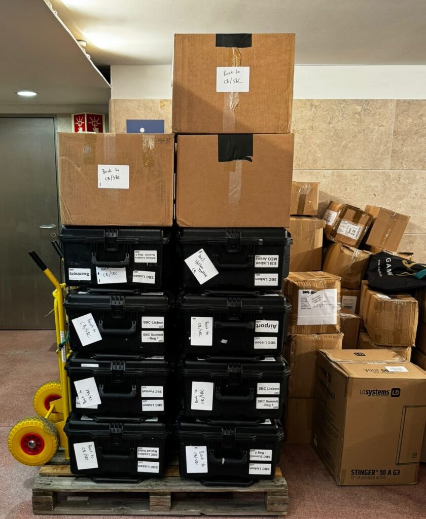 A stack of labelled black plastic cases and brown cardboard boxes on a wooden pallet in a back-of-house storage area, with additional boxes piled to the side and a yellow sack truck leaning against the stack.
