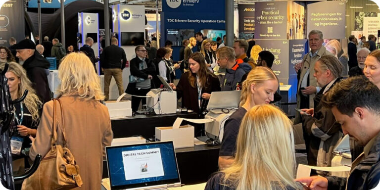 Busy conference registration desk with staff checking in attendees on laptops at the Digital Tech Summit, with crowded exhibition hall and booths visible in the background. late event registrations