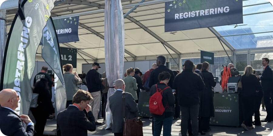 Attendees queuing at an outdoor event registration area under a marquee, with large 'Registrering' signs overhead and on-site badge printing equipment visible